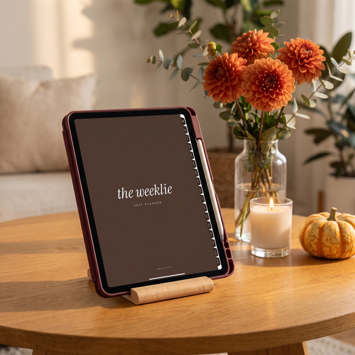 iPad in a burgundy case on a wood stand showing the 2027 digital planner cover with dahlias and a candle