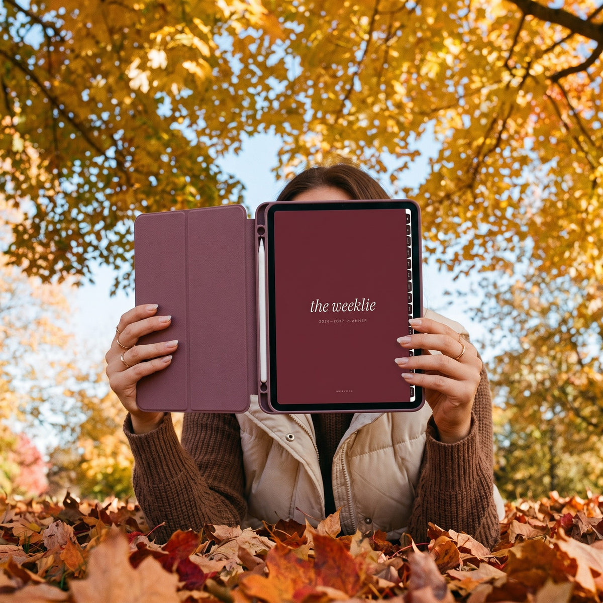 Lying in fall leaves holding up an iPad with the 2026-2027 Weeklie planner cherry cover