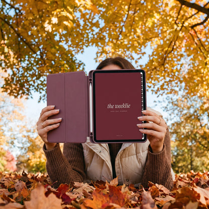 Lying in fall leaves holding up an iPad with the 2026-2027 Weeklie planner cherry cover