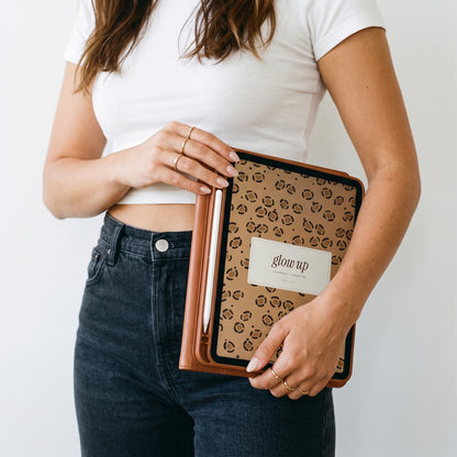 Woman in a white tee holding an iPad with the Glow-Up Journal leopard cover at her hip